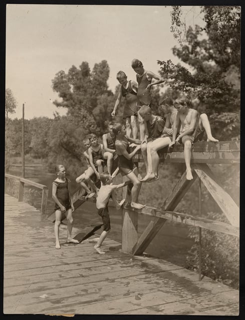 Lightly sepia-toned photograph of eleven young boys, wet from swimming, in bathing suits climbing a wooden ledge on a bridge over a creek. In the background, the creek flows underneath the bridge, surrounded by trees.